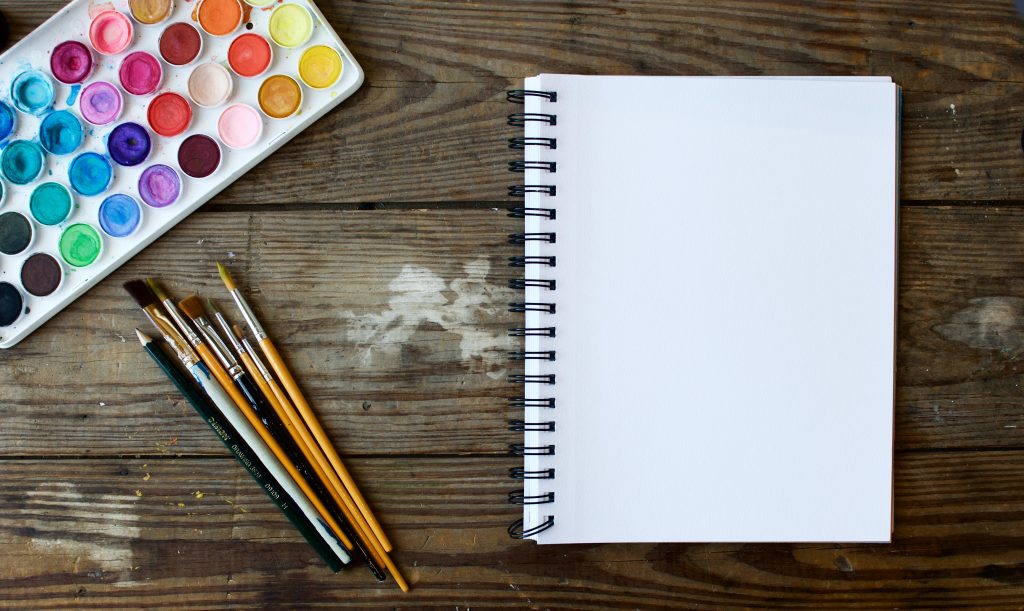 A desk with watercolors, paintbrushes, pencils, and a notebook perfect for creative nonfiction.