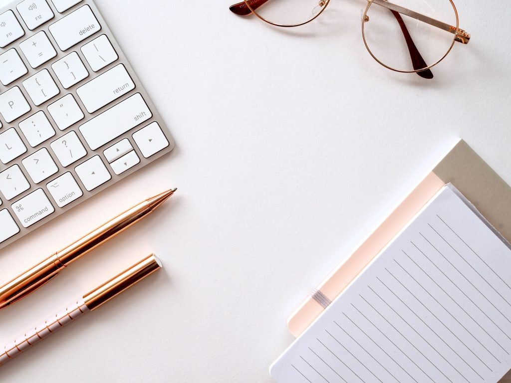 A desktop with a keyboard, glasses, writing utensils, and a notebook - everything needed to write guest posts.