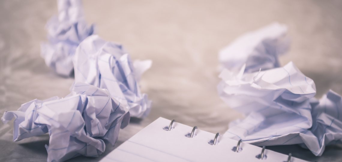 A blank notebook sits in the foreground, with crumpled pieces of paper balled up in the background from a reluctant writer with a serious case of writer's block.