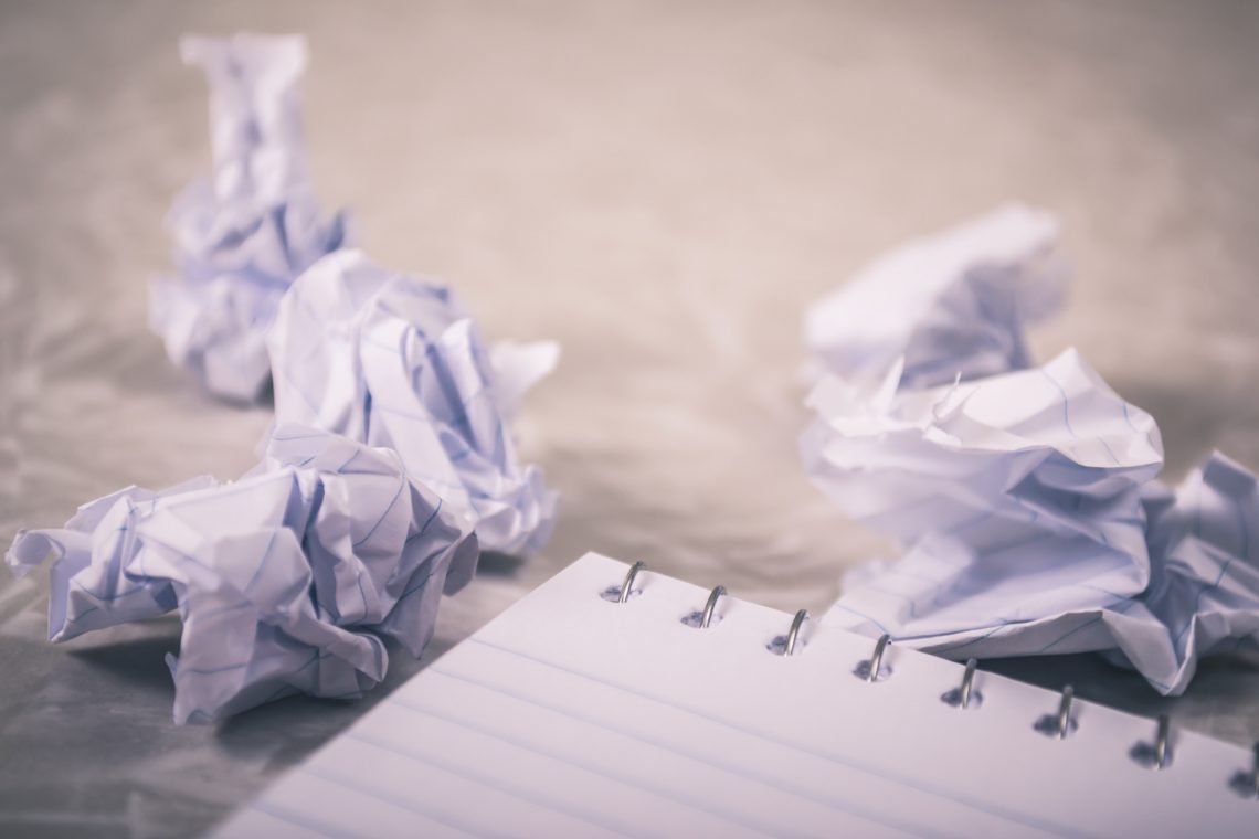 A blank notebook sits in the foreground, with crumpled pieces of paper balled up in the background from a reluctant writer with a serious case of writer's block.