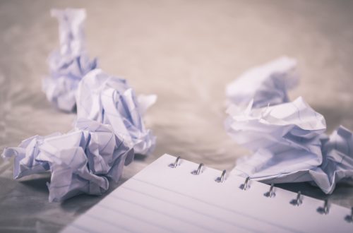 A blank notebook sits in the foreground, with crumpled pieces of paper balled up in the background from a reluctant writer with a serious case of writer's block.