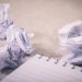 A blank notebook sits in the foreground, with crumpled pieces of paper balled up in the background from a reluctant writer with a serious case of writer's block.