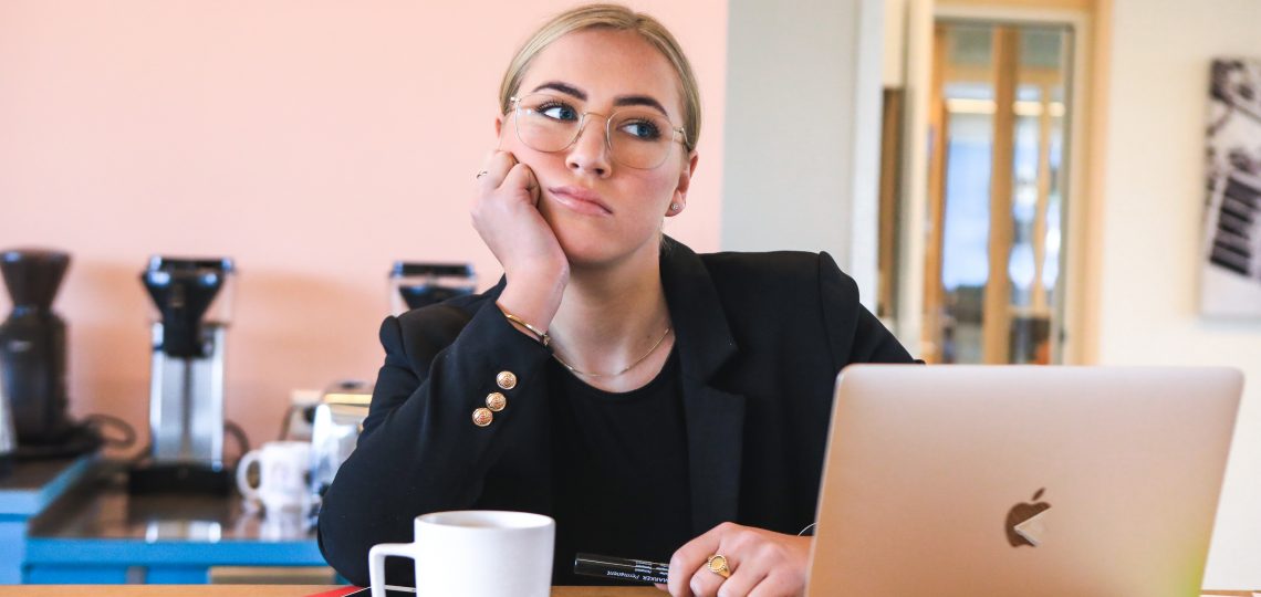 A woman sits at her desk with a laptop and a cup of coffee, hand on her chin. She's staring off in the distance, thinking "how do I stop procrastinating?"