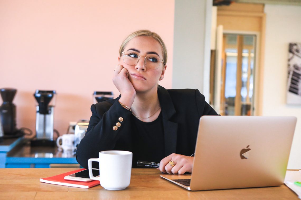 A woman sits at her desk with a laptop and a cup of coffee, hand on her chin. She's staring off in the distance, thinking "how do I stop procrastinating?"