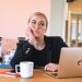 A woman sits at her desk with a laptop and a cup of coffee, hand on her chin. She's staring off in the distance, thinking "how do I stop procrastinating?"