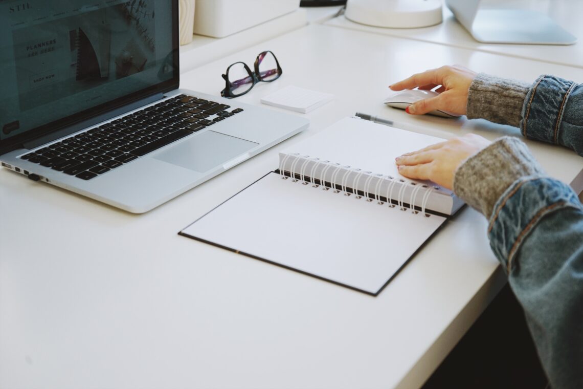 An author sitting down at her desk to write with a notebook, laptop, external mouse, and a pair of glasses sitting in front of her.