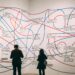 A man and a woman stand in front of a white wall full of phrases, with blue and red lines drawn between them, connecting them this way and that.