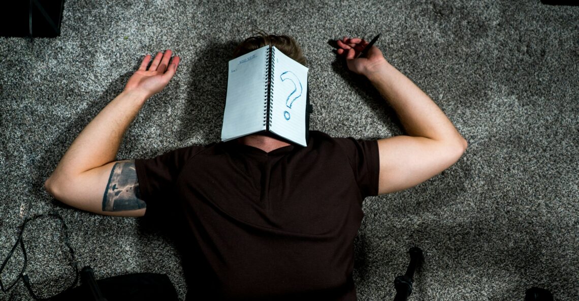 A man lies on the ground with a notebook over his face. One the notebook is a large question mark.