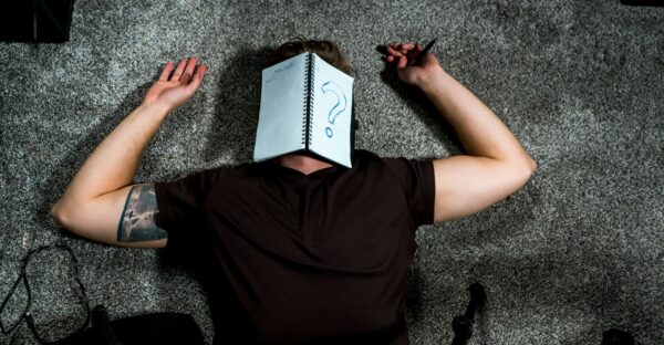 A man lies on the ground with a notebook over his face. One the notebook is a large question mark.