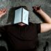 A man lies on the ground with a notebook over his face. One the notebook is a large question mark.